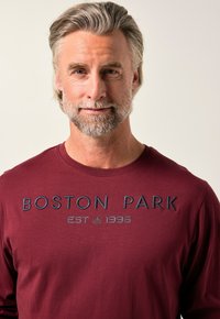 Middelbare man met grijs haar en baard, gekleed in een bordeauxrood shirt met de tekst "BOSTON PARK EST 1996" op de borst, neutrale achtergrond.