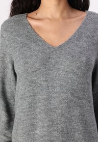 Pull en laine gris avec col en V, manches longues et texture douce. Présente une légère peluche et un détail minimaliste de couture au niveau du col.