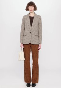 Beige porkkanapiste blazer, jossa on napitus, yhdistetty ruskeisiin mokkanahkaflare-housuihin. Malli pitää pientä valkoista käsilaukkua ja hänellä on mustat kengät.