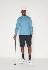 Lys blå hettegenser med glidelås, svarte shorts, hvit caps, holder en golfkølle. Joggeskoene er hvite med svarte detaljer og sportsstrømper.