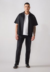 Μαύρο κοντομάνικο πουκάμισο με κουμπιά πάνω από ένα λευκό t-shirt, συνδυασμένο με σκούρα τζιν και μαύρα sneakers, με κομψό και μινιμαλιστικό σχέδιο.