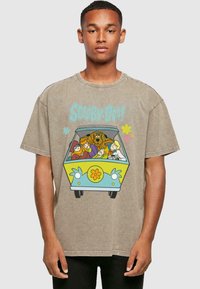 Camiseta de algodón color beige con un impreso colorido de "Scooby-Doo!" que muestra a los personajes en una furgoneta, con un diseño de flores amarillas en el vehículo.
