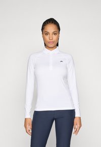 Frau trägt ein weißes Langarm-Polohemd mit einem kleinen schwarzen Logo auf der Brust und eine eng anliegende marineblaue Hose, stehend vor einem einfarbigen Hintergrund.