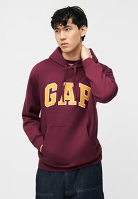 Νέος άνδρας με μπορντό φούτερ GAP με κίτρινα γράμματα, το ένα χέρι στην τσέπη, το άλλο αγγίζοντας τον λαιμό του, στέκεται μπροστά σε ένα λευκό φόντο.