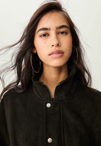 Jeune femme aux longs cheveux noirs et grandes boucles d'oreilles en anneau porte une veste noire à col haut sur un fond clair uniformément.