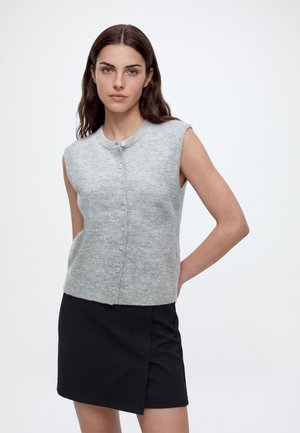 Kvinde med langt brunt hår iført en ærmeløs grå knappet sweater og et sort asymmetrisk skørt, står med hænderne bag ryggen.