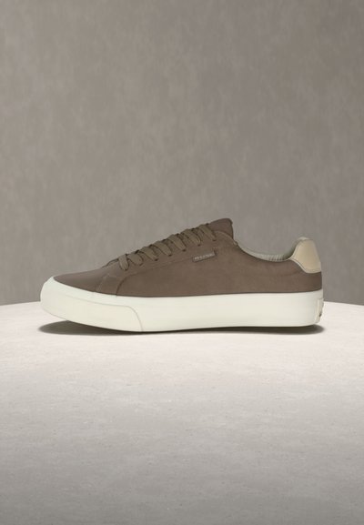 Baskets basses marron avec semelle blanche et accent au talon, lacets sur le côté et petite étiquette métallique de la marque sur un fond neutre.