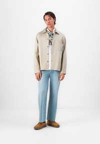 Beige cotton shirt jacket with front pocket, brown buttons, and relaxed fit, paired with light blue denim jeans and brown shoes. White background.