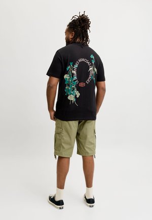 Homme avec dreadlocks portant un t-shirt noir avec un motif floral et un texte, un short cargo olive, des chaussettes blanches et des baskets noires, debout, de dos.