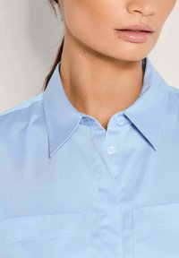 Vrouw in een lichtblauw collared shirt met knopen, met een close-up van haar nek en lippen.