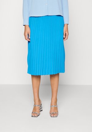 Vrouw draagt een felblauwe geplooide midirok, lichtblauwe top met knopen en zwart-witte open teen hakken met ruitpatroon.