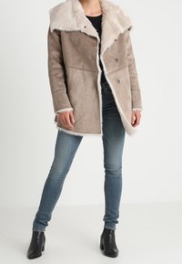 Taupe faux suede bunda s velkým límcem z umělé kožešiny, dvouřadým zapínáním a dlouhými rukávy, spojená s modrými skinny džíny a kotníkovými botami.