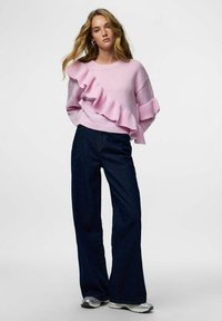 Jasnoróżowy dzianinowy sweter z falbanką po lewej stronie, zestawiony z ciemnymi dzwonowatymi dżinsami i srebrnymi trampkami.