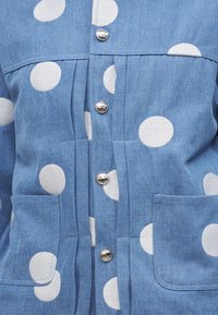 Blue denim jacket with large white polka dots, metallic snap buttons down center, and two front patch pockets.