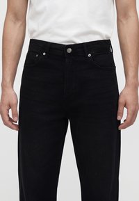 Vue de face d’une personne portant un jean noir et une chemise blanche, les mains détendues le long du corps sur un fond uni.