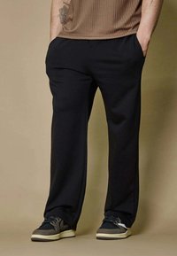 Homme portant un pantalon noir ample et des baskets noir et blanc, se tenant avec les mains dans les poches devant un fond beige.