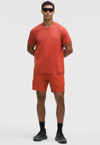 Punainen urheilupaita lyhyillä hihoilla, yhdistetty samaan punaiseen shortseihin. Materiaalina kevyt kangas, asuun kuuluu mustat lenkkarit ja aurinkolasit.