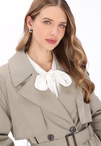 Beige trenchcoat met grote kraag, dubbele knopenrij en een bijpassende riem. Witte blouse met een strikdetail steekt eronder uit.