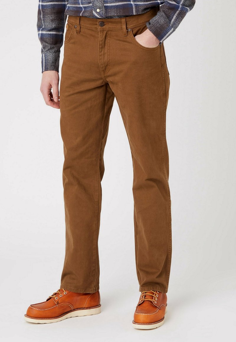 Man med bruna rakbenade byxor, beige läder snörkängor och en blårutigt långärmad skjorta, hand i fickan.