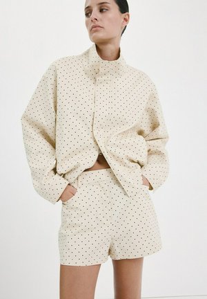 Mujer de cabello oscuro con chaqueta beige de lunares grandes y pantalones cortos a juego, con las manos en los bolsillos, mirando hacia un lado sobre un fondo blanco.