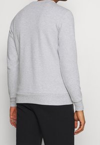 Hombre vestido con una sudadera de manga larga gris claro y pantalones negros, visto desde la espalda contra un fondo claro y liso.