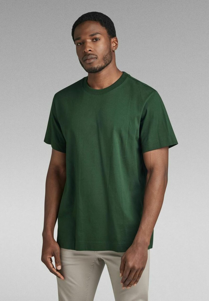 T-shirt verde in cotone con maniche corte, scollatura rotonda e una texture liscia. Indossato con pantaloni di colore chiaro. Design semplice, colore uniforme.