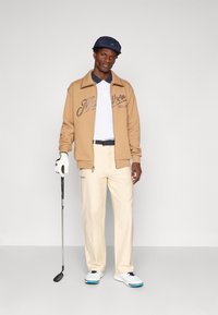 Brūns zip-up jaciņš ar "Marlon Golf" uzrakstu, balta polo krekls, krēmkrāsas bikses un baltas golfu kurpes ar ziliem akcentiem. Tur rokās golfa nūju.