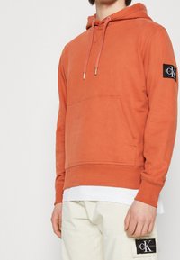 Orange bomullshoodie med en ficka fram, ribbat avslut och muddar. Har en svart logotyp-patch på höger ärm. Avslappnad design.