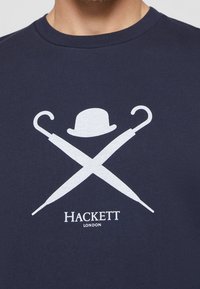 T-shirt in cotone navy con una grafica bianca di ombrelli incrociati e un cappello bowler sopra il nome del marchio "Hackett London" in testo bianco.