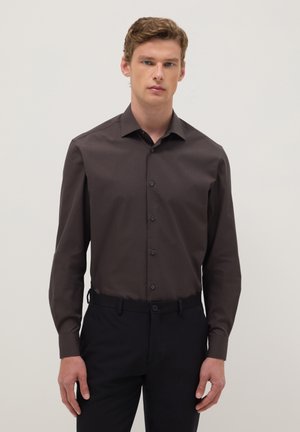 Jeune homme portant une chemise brune à boutons et un pantalon noir, debout devant un fond clair uni, faisant face à l'avant.