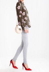 Blusa de seda verde olivo con estampado floral, jeans ajustados gris claro, tacones de aguja rojos y un pequeño bolso de hombro redondo color crema.