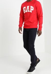 Felpa rossa con logo "GAP" bianco, abbinata a pantaloni slim-fit blu navy e sneakers nere. Design semplice e casual, con una vestibilità confortevole.