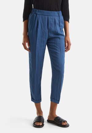 Nainen, jolla on yllään siniset kapenevat housut, joissa on käännetyt lahkeet, musta pitkähiha toppi ja mustat slip-on sandaalit, seisoo yksiväristä taustaa vasten.