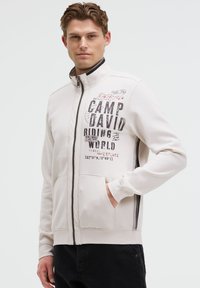 Helle beige Sweatjacke mit Reißverschluss, die grafischen Text enthält, zwei vordere Taschen und einen kontrastierenden dunklen Saum entlang des Reißverschlusses und der Handgelenke hat.