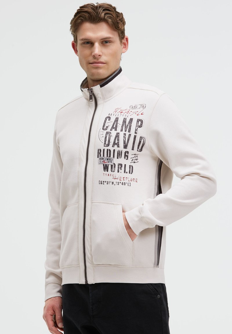 Helle beige Sweatjacke mit Reißverschluss, die grafischen Text enthält, zwei vordere Taschen und einen kontrastierenden dunklen Saum entlang des Reißverschlusses und der Handgelenke hat.