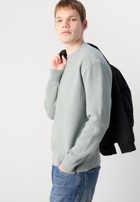Hellgrüner, gerippter Pullover mit langen Ärmeln, rundem Ausschnitt und strukturiertem Stoff; kombiniert mit einer schwarzen Bomberjacke, die über die Schulter gelegt ist.