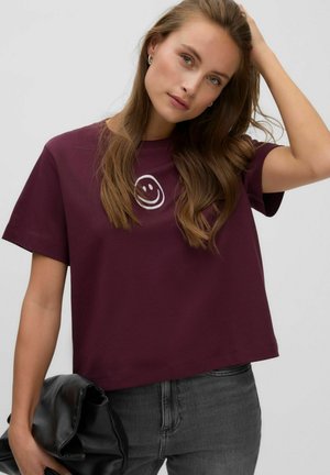 Kvinde med langt brunt hår iført en vinrød cropped t-shirt med et hvidt smiley ansigt og grå jeans, holder en sammenfoldet sort jakke.