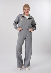 Jeune femme en sweat-shirt gris clair à fermeture éclair et pantalon large assorti ajustant l'ourlet, debout devant un fond uni.