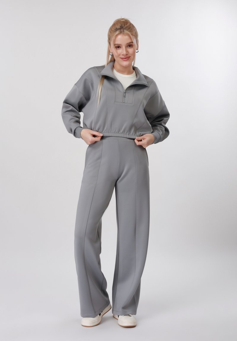 Jeune femme en sweat-shirt gris clair à fermeture éclair et pantalon large assorti ajustant l'ourlet, debout devant un fond uni.