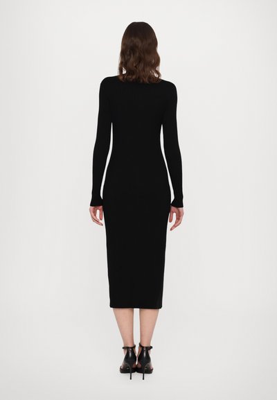 Vestido midi negro de canalé con mangas largas, silueta ajustada y cuello redondo. Llevado con tacones negros de pulsera al tobillo.
