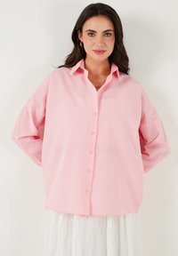 Light pink oversized button-up shirt with a pointed collar and long sleeves, featuring a smooth cotton material and simple button detailing.