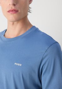 Modrá bavlnená tričko s okrúhlym výstrihom a krátkymi rukávmi, s malým bielym logom "HUGO" na hrudi. Hladká textúra.