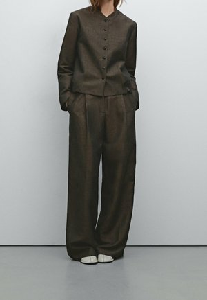 Person wearing loose dark brown buttoned blazer and wide-legged trousers with white shoes, hands in pockets, against plain light background.