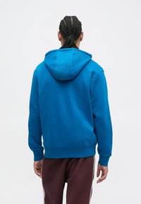 Sudadera de algodón azul con capucha ajustable, puños acanalados y dobladillo. Combinada con pantalones burdeos con franjas laterales. Vista trasera.