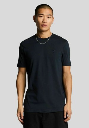 Ung mand med kort hår iført en almindelig sort t-shirt, sølvkæde og ringe, stående mod en grå baggrund.