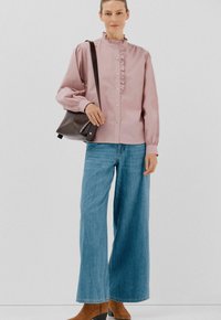 Donna che indossa una blusa rosa a balze, jeans blu a gamba larga, stivali alla caviglia marroni e porta una borsa a spalla marrone scuro.