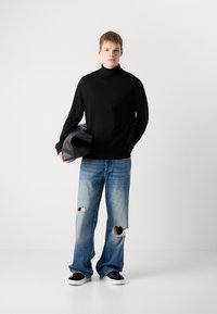 Fekete garbós pulóver, kék, kopott, bő szabású farmernadrág nagy lyukakkal, és fekete tornacipő. A modell egy fekete táskát tart.