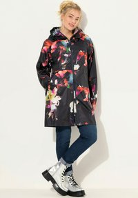 Regenjack gemaakt van glanzend zwart materiaal met een kleurrijk bloemmotief, ritsluiting, zakken en een capuchon; gedragen met blauwe jeans en zilveren laarzen.