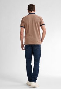 Brun pikétröja med en texturerad design, marinblå krage och ärmdekorationer, tillsammans med mörkblå jeans och ljusa sneakers.