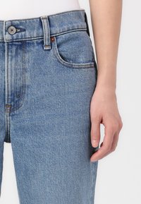 Jean en denim bleu clair avec une taille ajustée, un design à cinq poches, des coutures visibles et une texture légèrement délavée.
